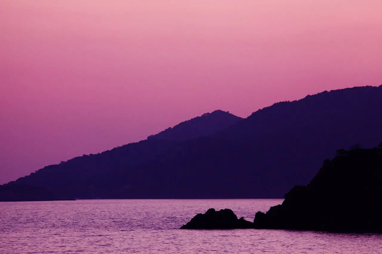 A tranquil seascape at sunset with calm water, silhouetted rocky shoreline, and rolling hills in the background under a pink and purple sky.