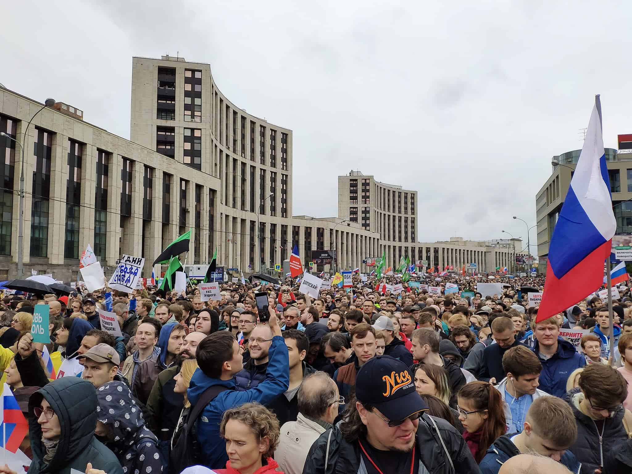 A large crowd of people gathers for a protest in a city, holding signs, banners, and flags, including a prominent Russian flag, with modern buildings in the background under a cloudy sky.