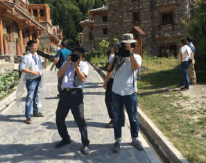 A group of people stands on a stone pathway in front of rustic buildings; two men in the center point cameras toward the viewer while others stand or walk in the background.
