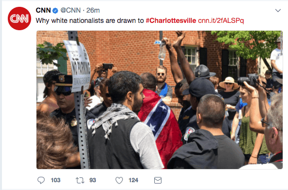A group of people, some wearing hats and uniforms, gather around a man draped in a Confederate flag during a protest in Charlottesville. Several are filming or taking photos, and a sign reading “NO PARKING” is visible.