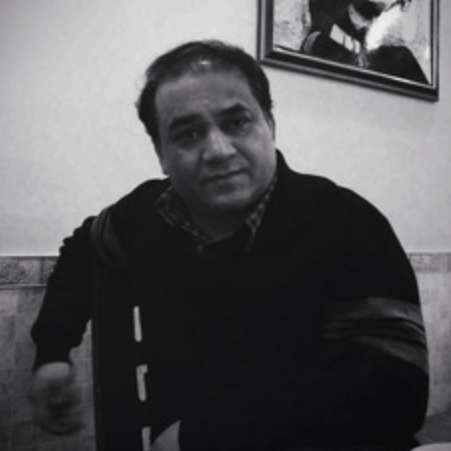 A man with short dark hair sits indoors, wearing a dark sweater over a plaid shirt. He looks toward the camera with a neutral expression. A framed picture hangs on the wall behind him. The image is in black and white.