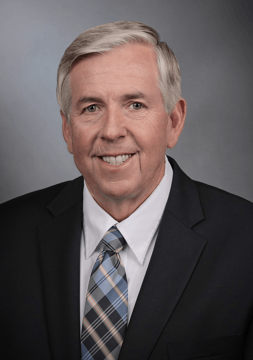 Smiling older man with short gray hair wearing a dark suit, white shirt, and a plaid tie, posed in front of a plain gray background.