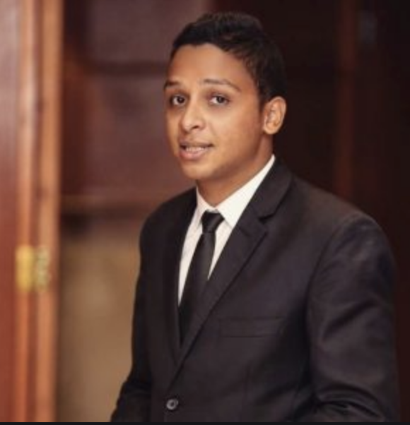 A young man in a black suit and tie stands indoors, looking at the camera and smiling softly. The background is out of focus with wooden tones.