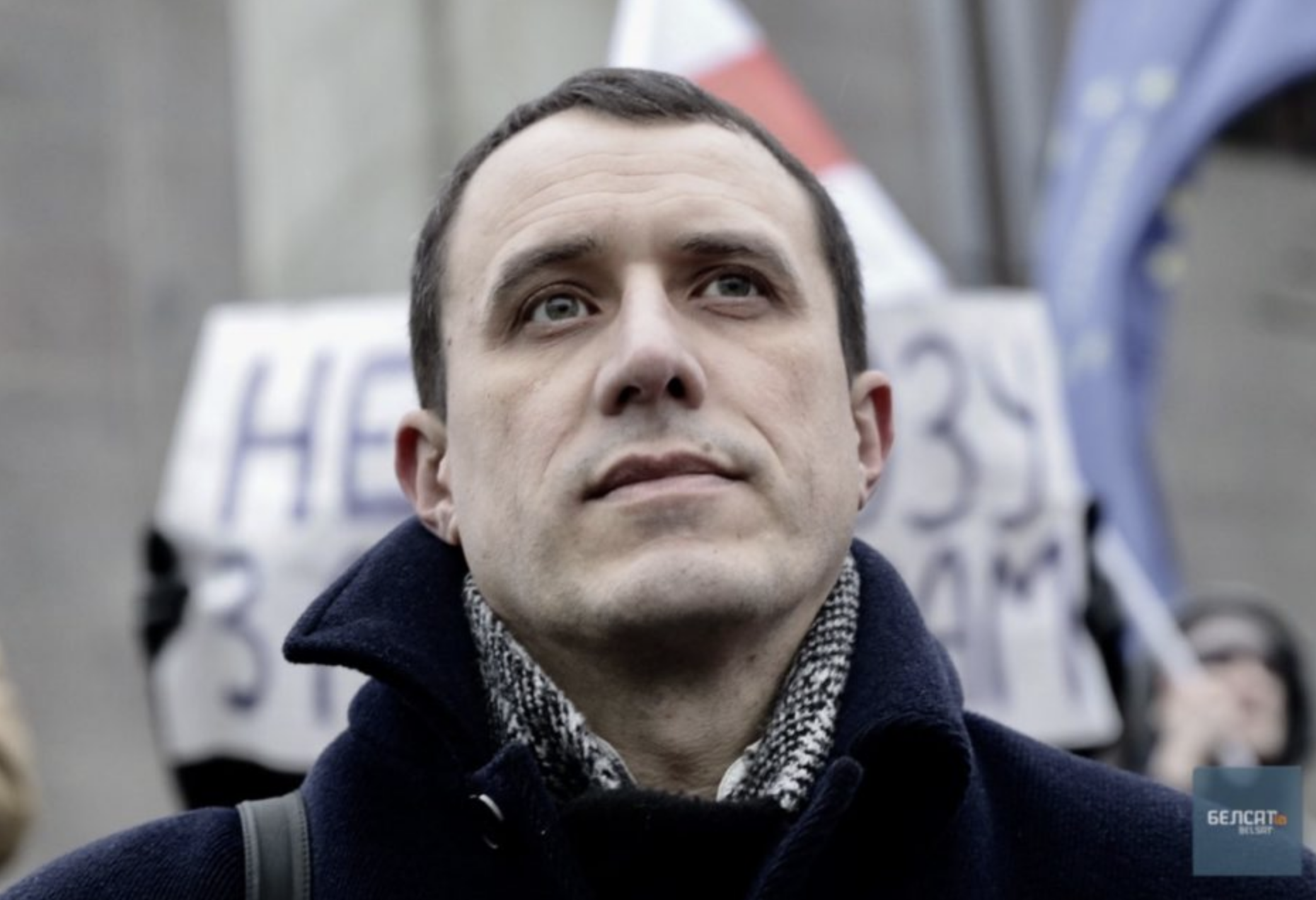 A man wearing a dark coat and scarf looks slightly upward with a serious expression. Blurred protesters and signs are visible in the background.