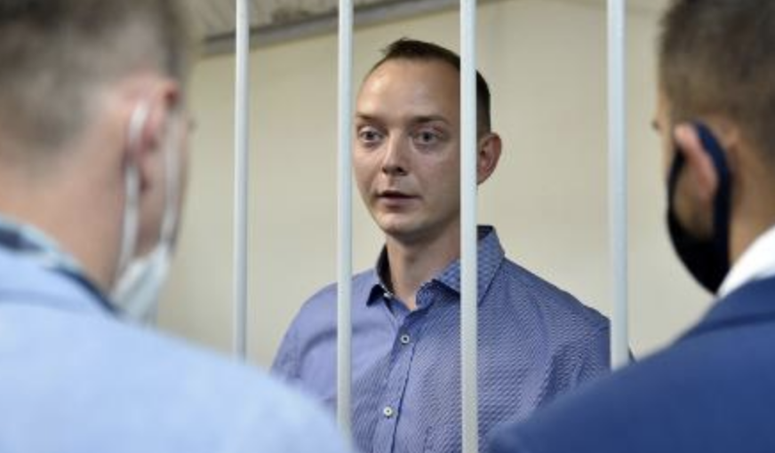A man in a blue shirt stands behind metal bars, facing two men in suits and masks who are standing on the other side of the bars. The setting appears to be an official or detention facility.