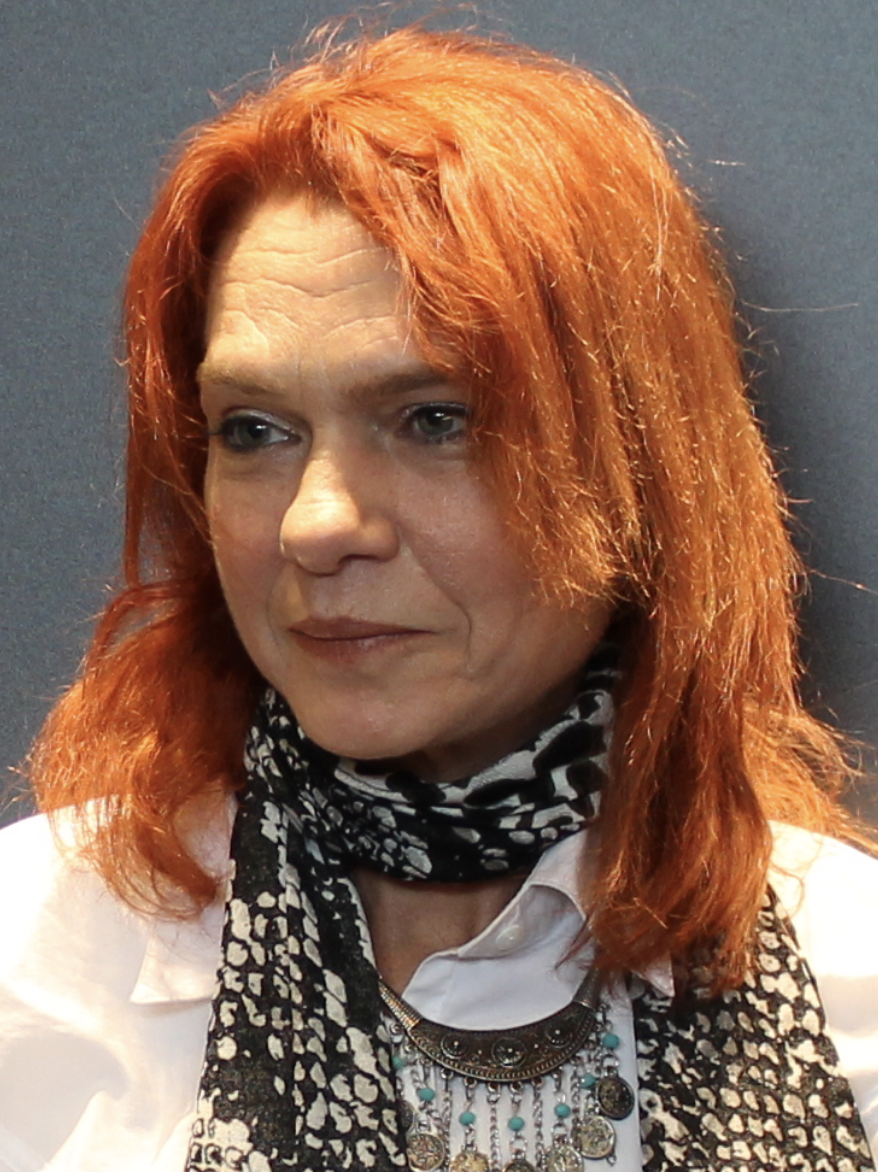 A person with shoulder-length red hair is wearing a white shirt, a patterned scarf, and a decorative necklace, standing against a plain gray background.