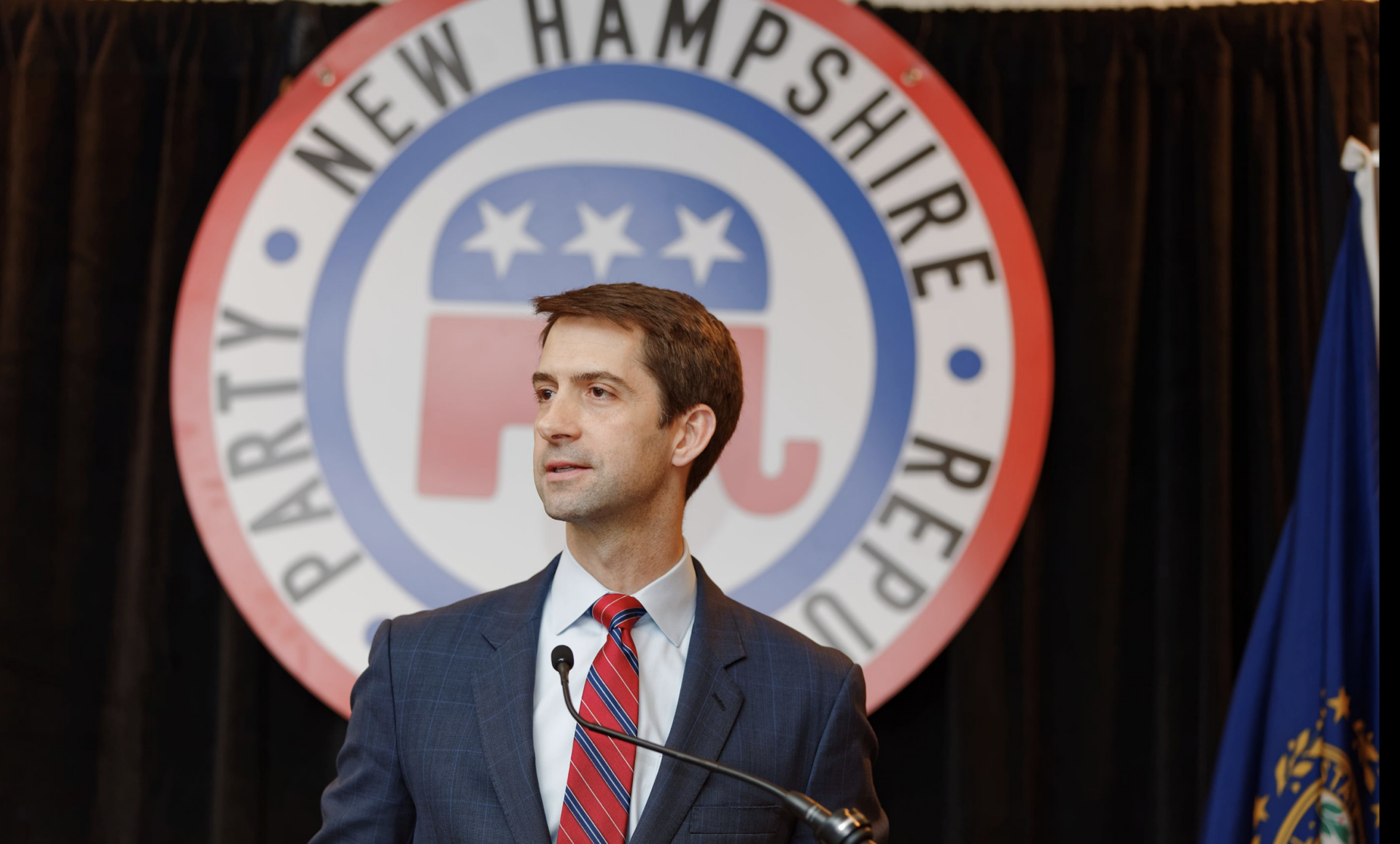 A man in a suit and red striped tie speaks at a podium in front of a large New Hampshire Republican Party sign featuring an elephant symbol.