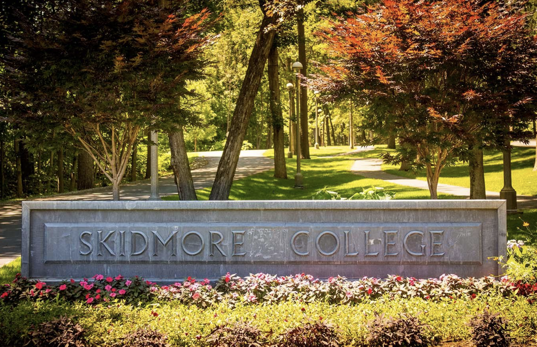 A stone sign reading Skidmore College stands amid colorful flowers and vibrant trees, with a sunlit campus pathway and greenery visible in the background.