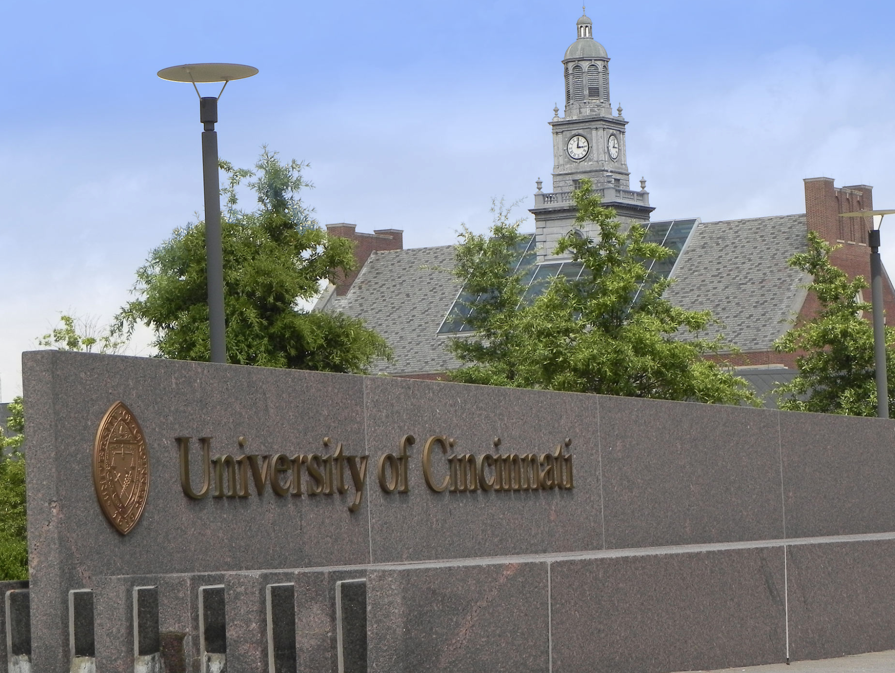 A stone sign with University of Cincinnati engraved on it stands in front of trees and a campus building with a clock tower under a partly cloudy sky.