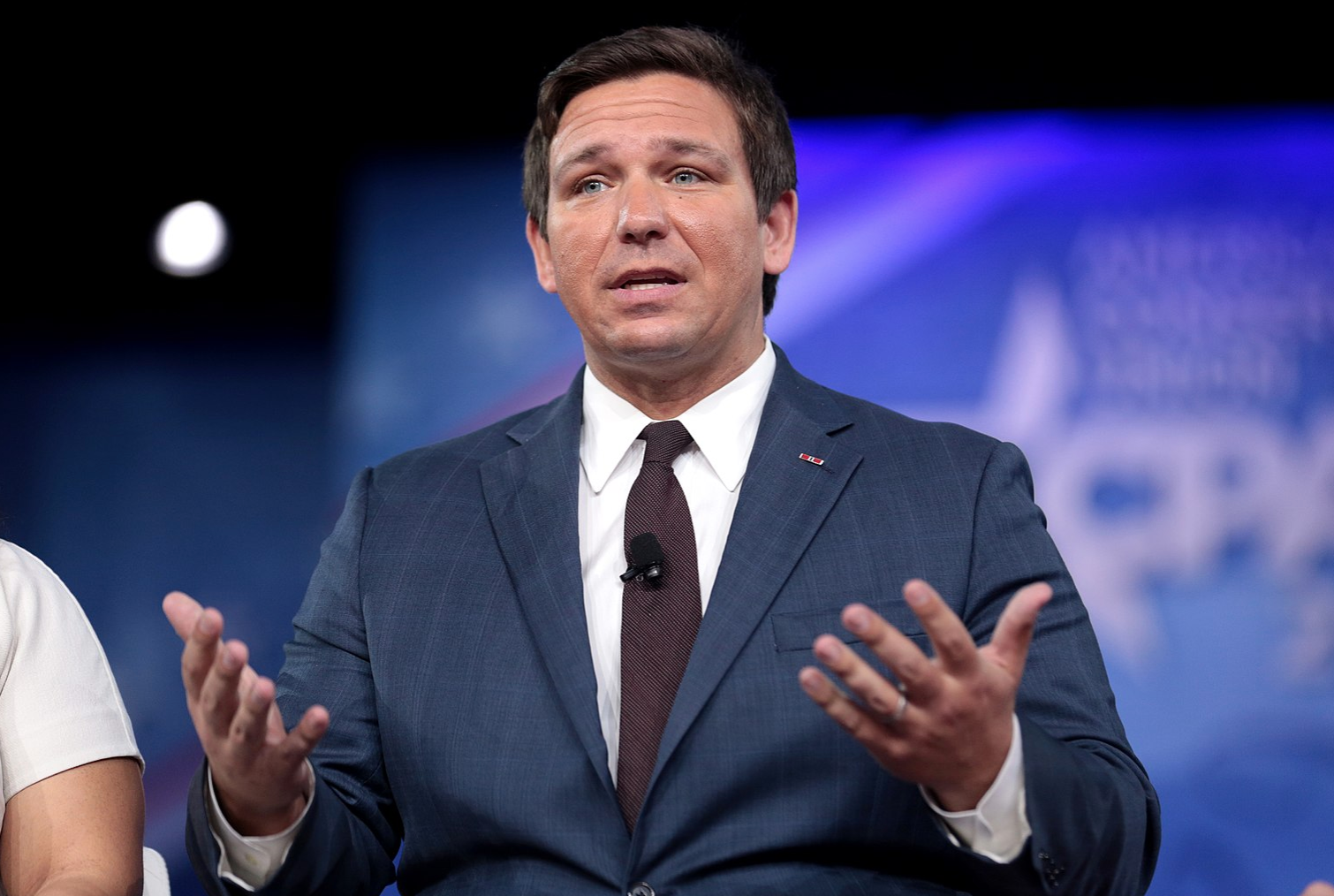 A man in a blue suit and dark tie is speaking on stage with his hands raised. The background is blue with blurred text and graphics.