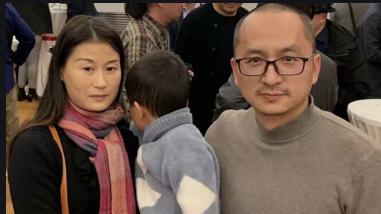 A woman and a man stand together at an indoor event, with the man holding a young child facing away from the camera. The woman wears a scarf, and the man wears glasses and a gray sweater. Other people are in the background.