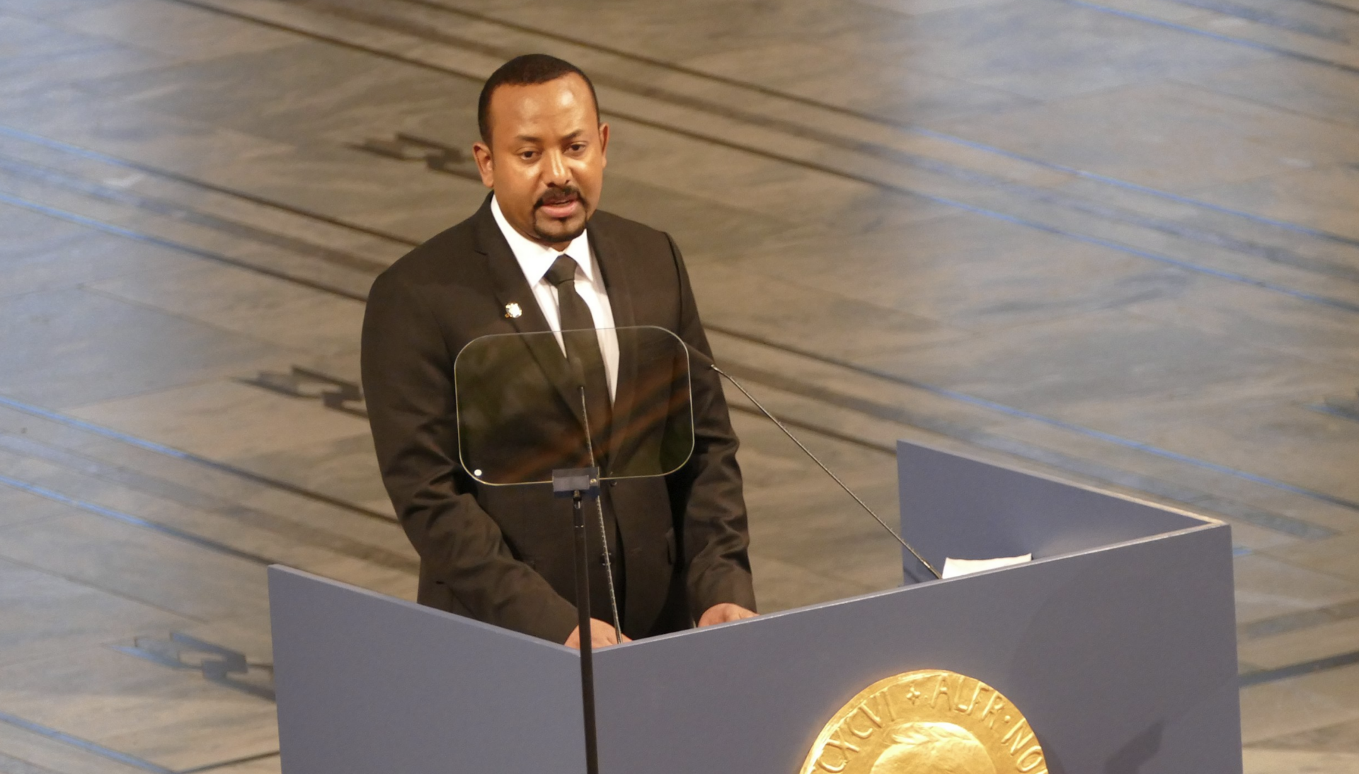 A man in a black suit and tie stands at a podium with a gold emblem, speaking into a microphone, with a clear teleprompter in front of him. The background is a large, light-colored stone floor.