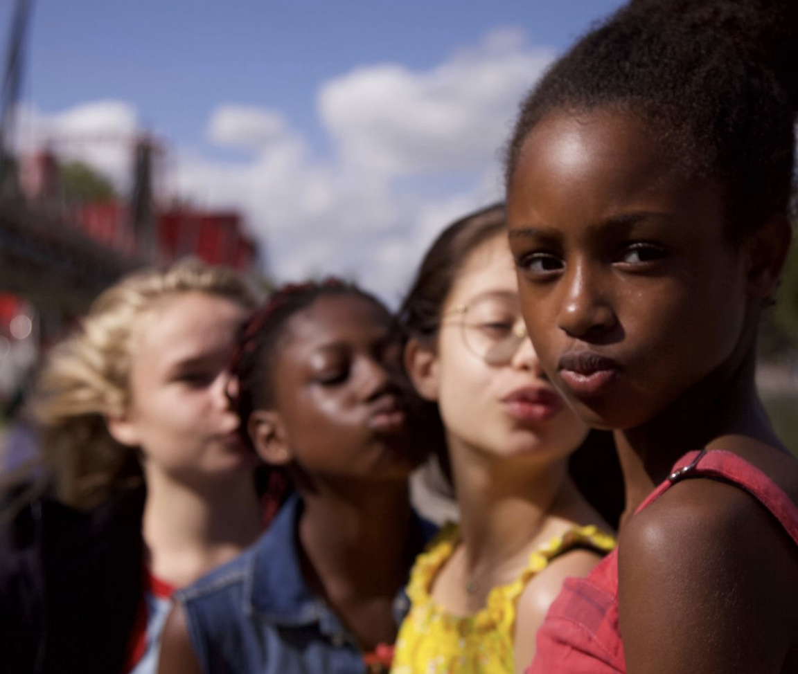 Four girls pose together outdoors on a sunny day; three are blurred in the background, while one girl in a red top stands in sharp focus at the front, looking directly at the camera and making a pouty face.