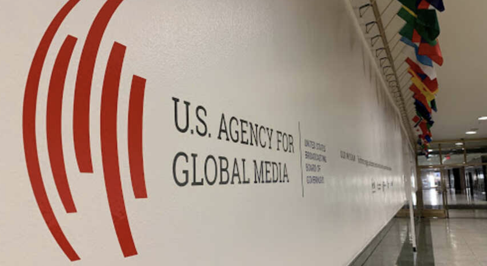 A hallway with a wall displaying the U.S. Agency for Global Media logo and name in large text; a row of international flags hangs from the ceiling on the right.