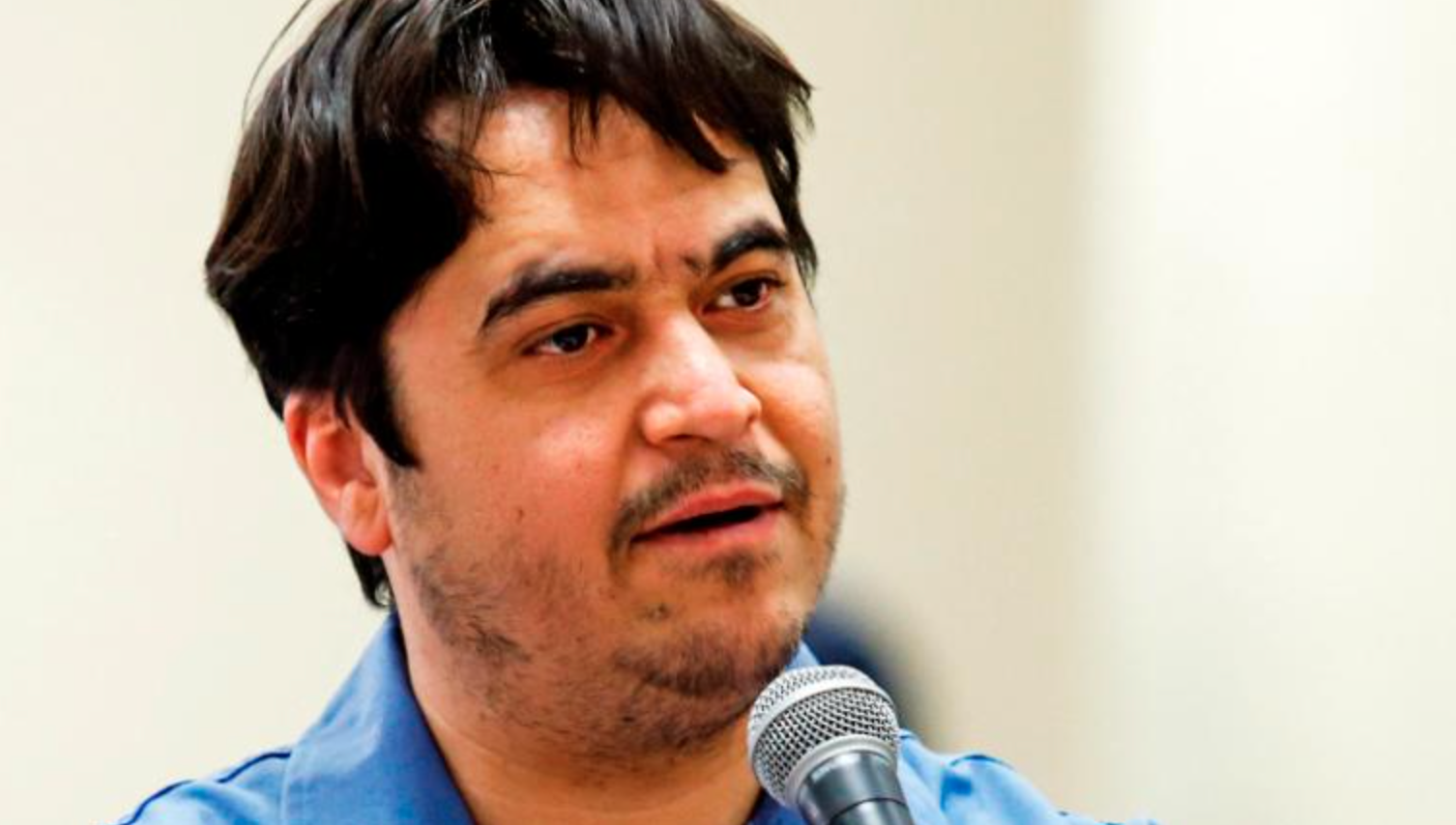 A man with short dark hair and facial stubble speaks into a microphone. He is wearing a blue collared shirt and there is a plain light background behind him.