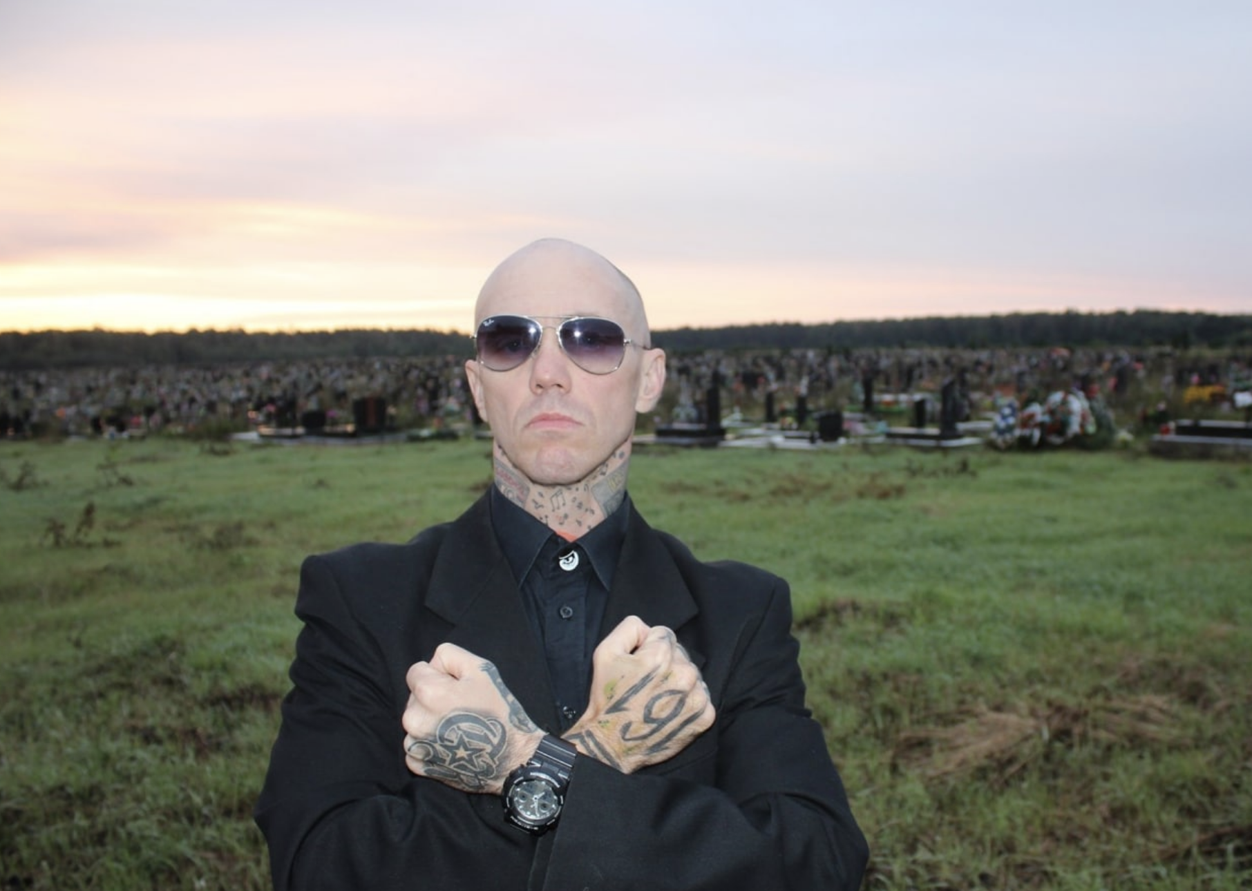 A bald man in sunglasses and a black suit stands in a grassy cemetery at sunset, arms crossed in front of his chest. He has visible tattoos on his hands and neck and wears a serious expression.