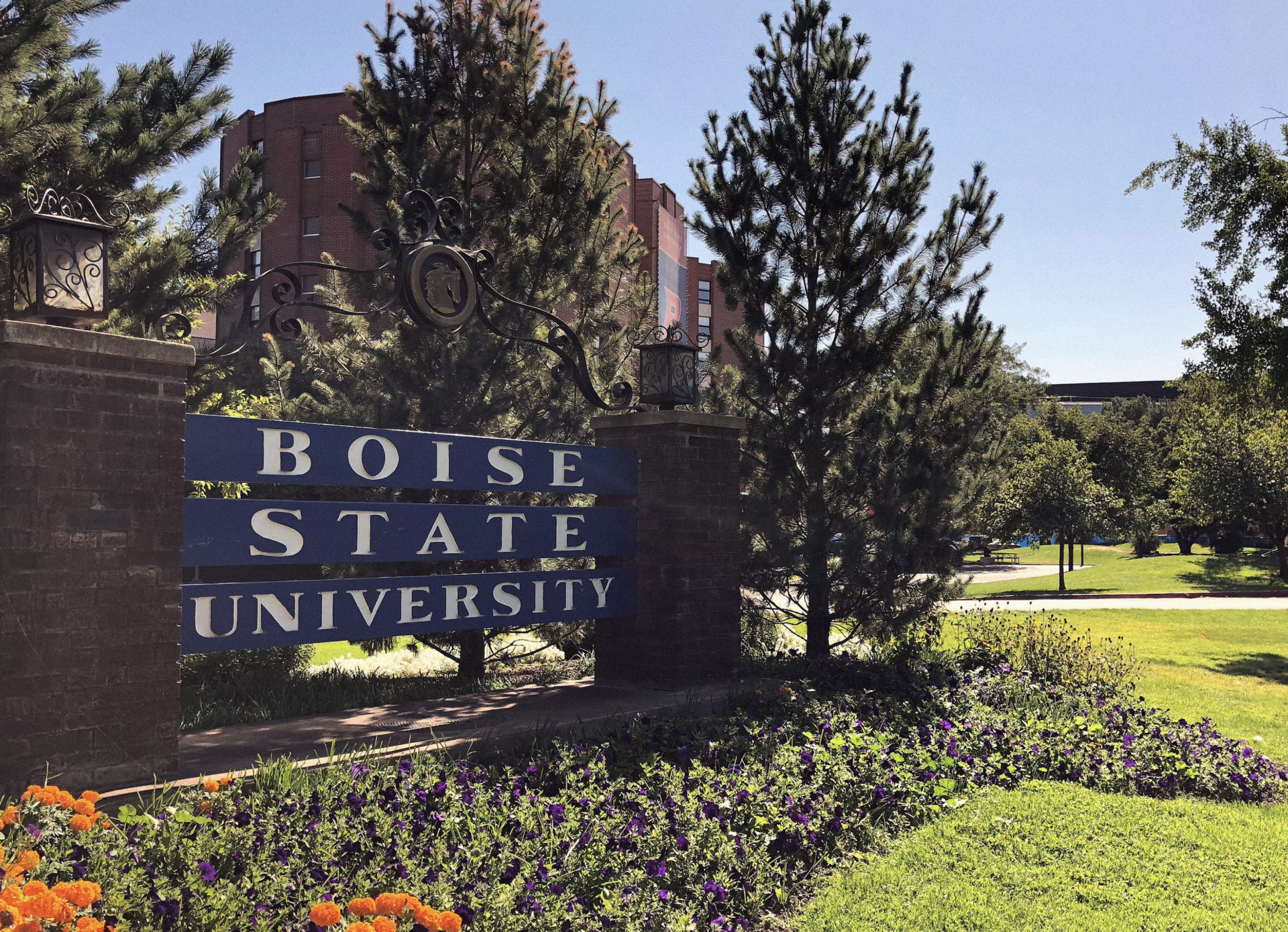 A blue and white sign reading Boise State University stands among flowers and pine trees, with a brick building and clear blue sky in the background.