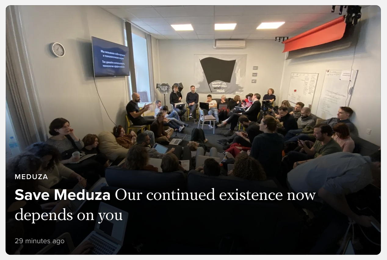 A large group of people sits closely together in a small, well-lit room, some with laptops and notepads, engaged in discussion. A sign on the wall reads Save Meduza. The atmosphere appears serious and focused.
