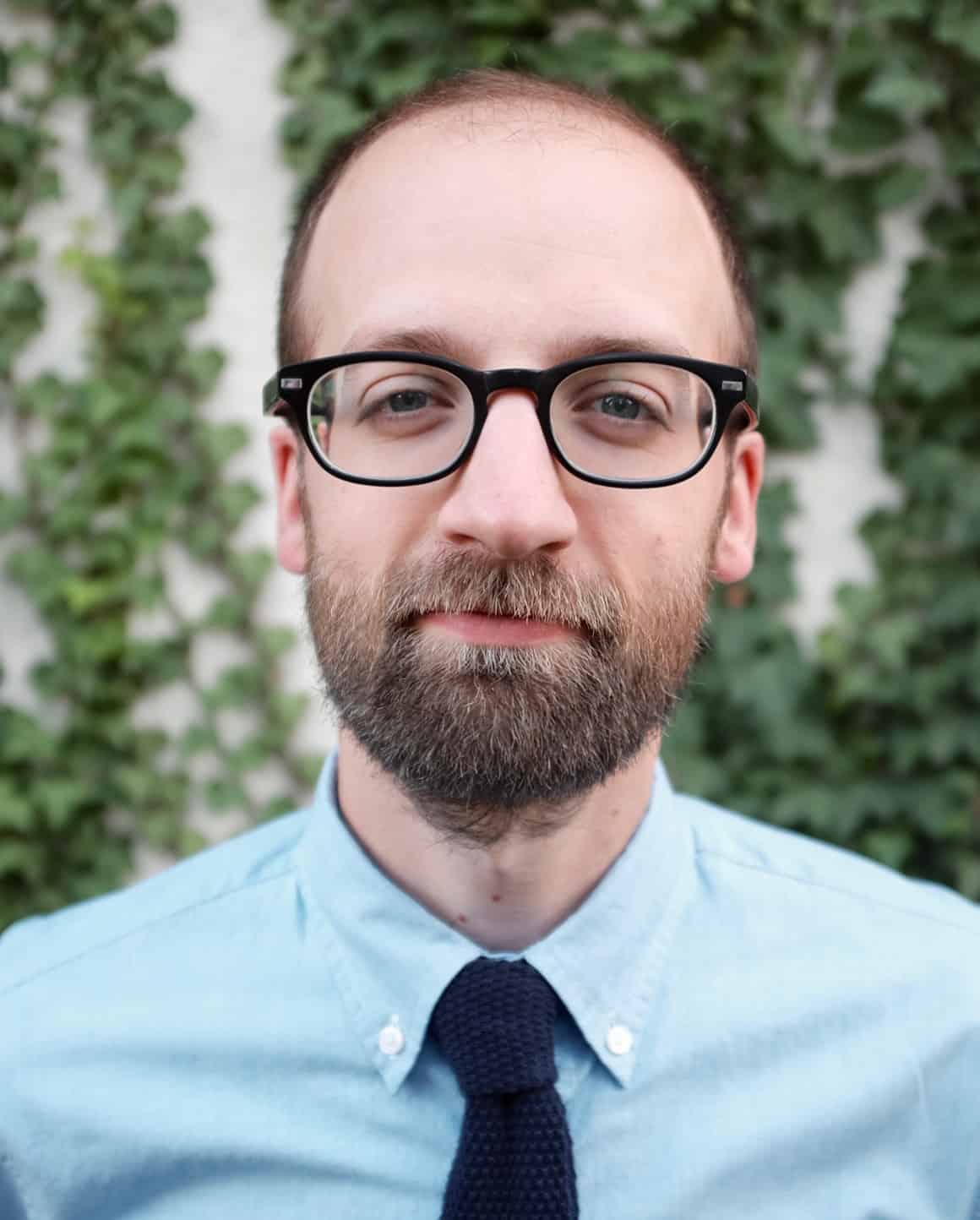 A man with short hair, a full beard, and black glasses is wearing a light blue collared shirt and a dark tie, standing in front of a wall covered with green ivy leaves.