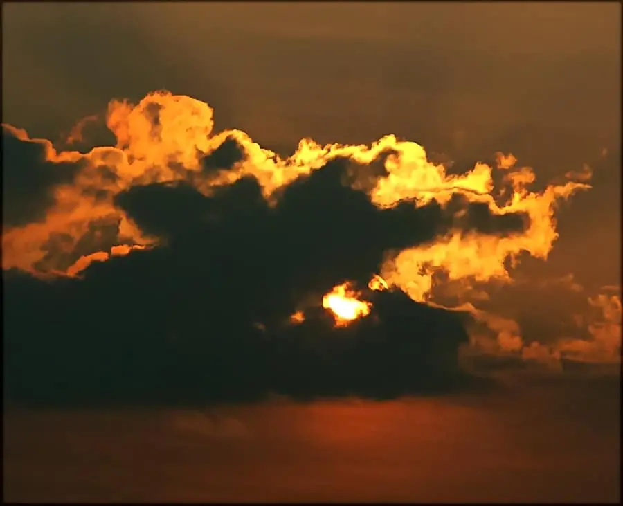 Dark clouds partially obscure a glowing orange sunset, with the sun peeking through the clouds and casting warm light across the sky.