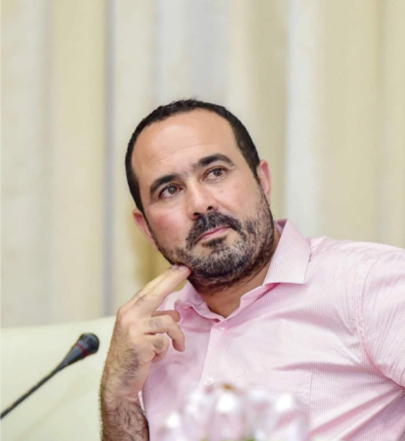 A man with short dark hair and a trimmed beard, wearing a pink shirt, sits thoughtfully with one hand touching his face. A microphone is visible in front of him, and the background is softly blurred.
