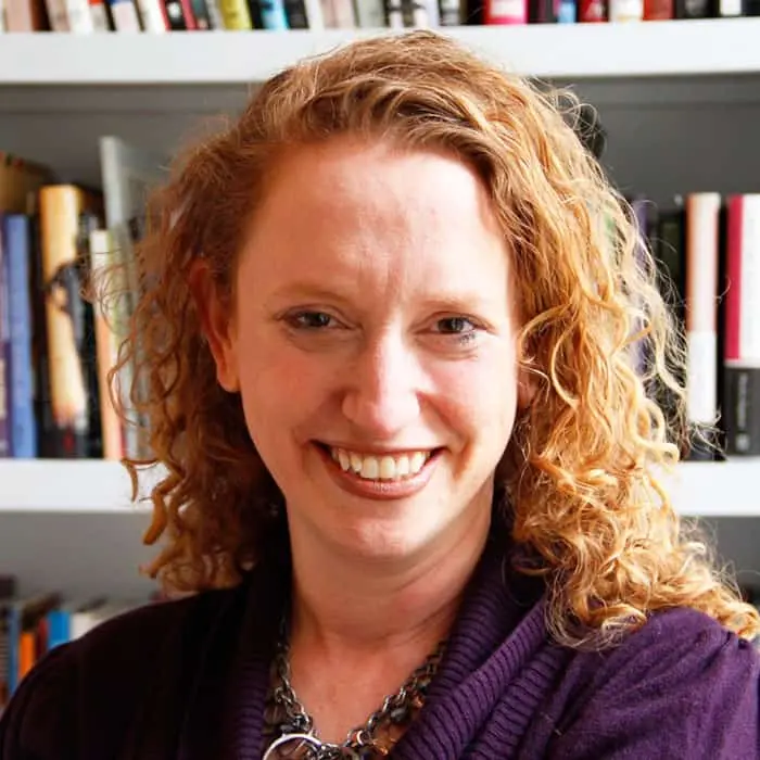 A woman with curly red hair smiles at the camera, wearing a dark purple sweater and a necklace, standing in front of a bookshelf filled with books.