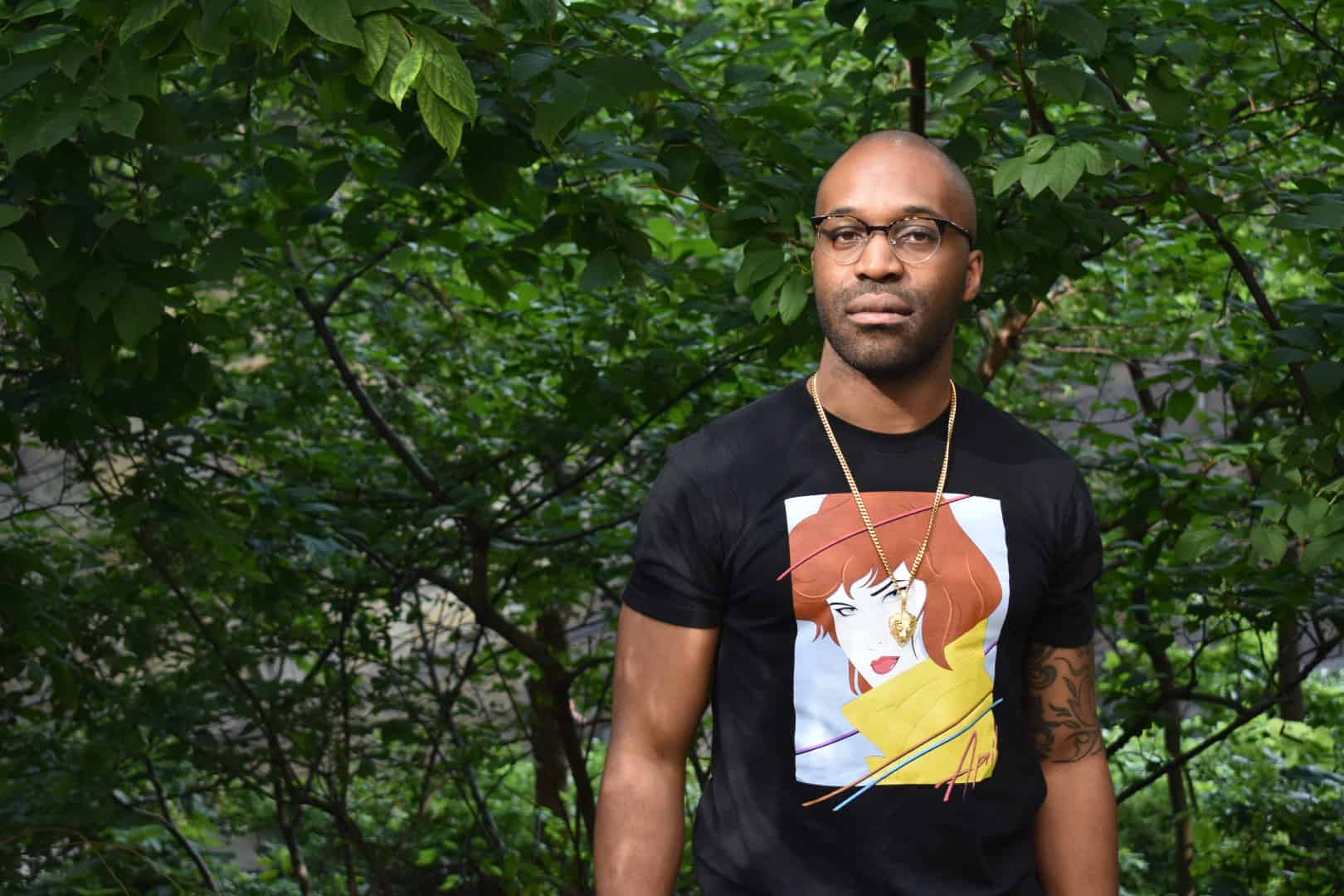 A man wearing glasses and a black T-shirt with a colorful illustration of a woman stands outdoors in front of dense green foliage, as if ready for a pen ten interview. He has a tattoo on his left arm and is wearing a gold necklace.