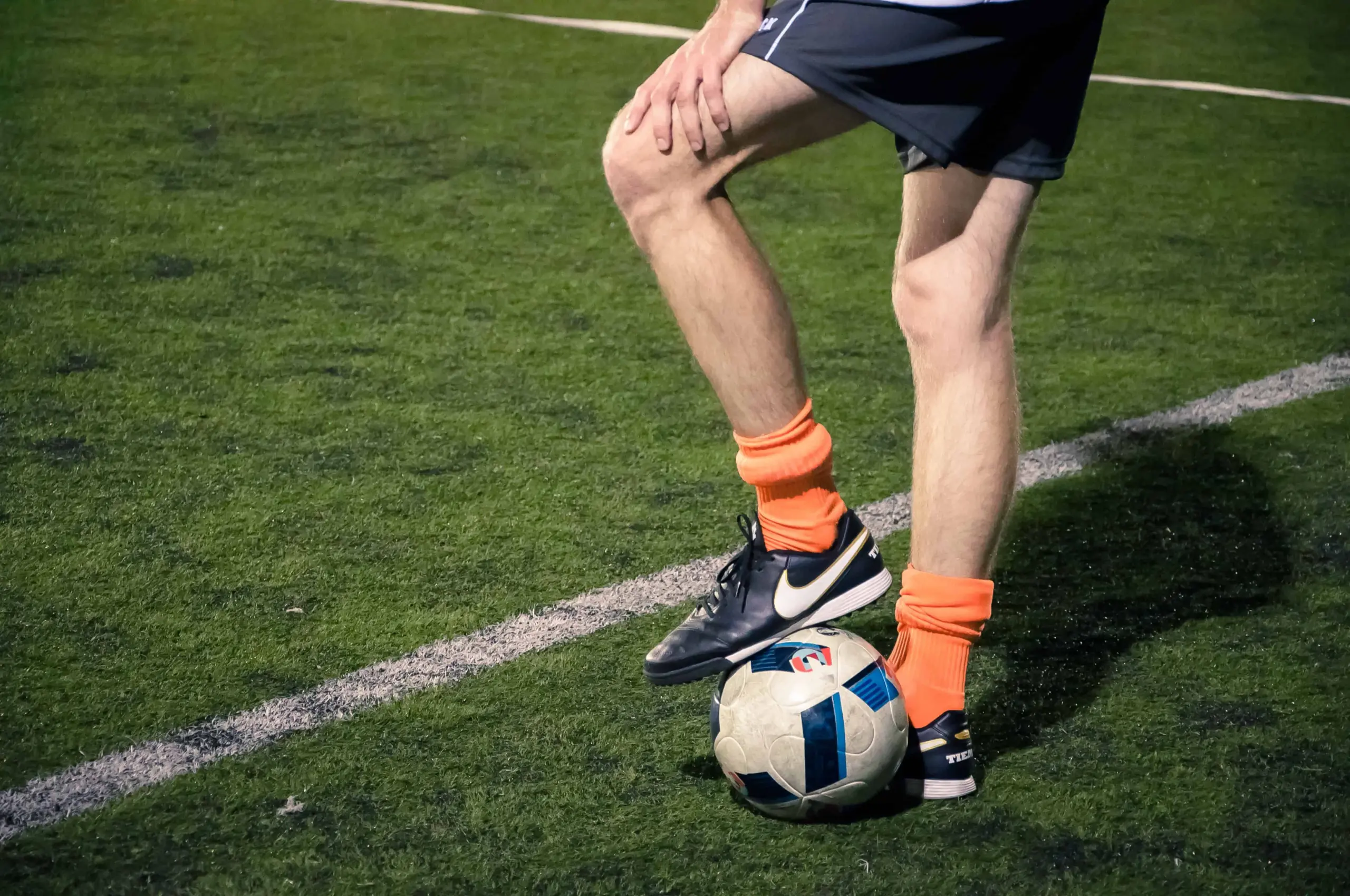 A soccer player wearing orange socks and black cleats stands on a green field, resting one foot on a soccer ball near a white sideline. Only the player’s lower body is visible.