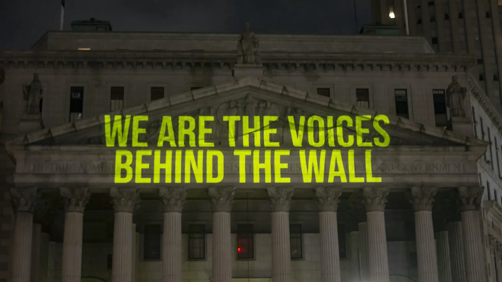 The words “we are the voices behind the wall” being projected in bright green on the building
