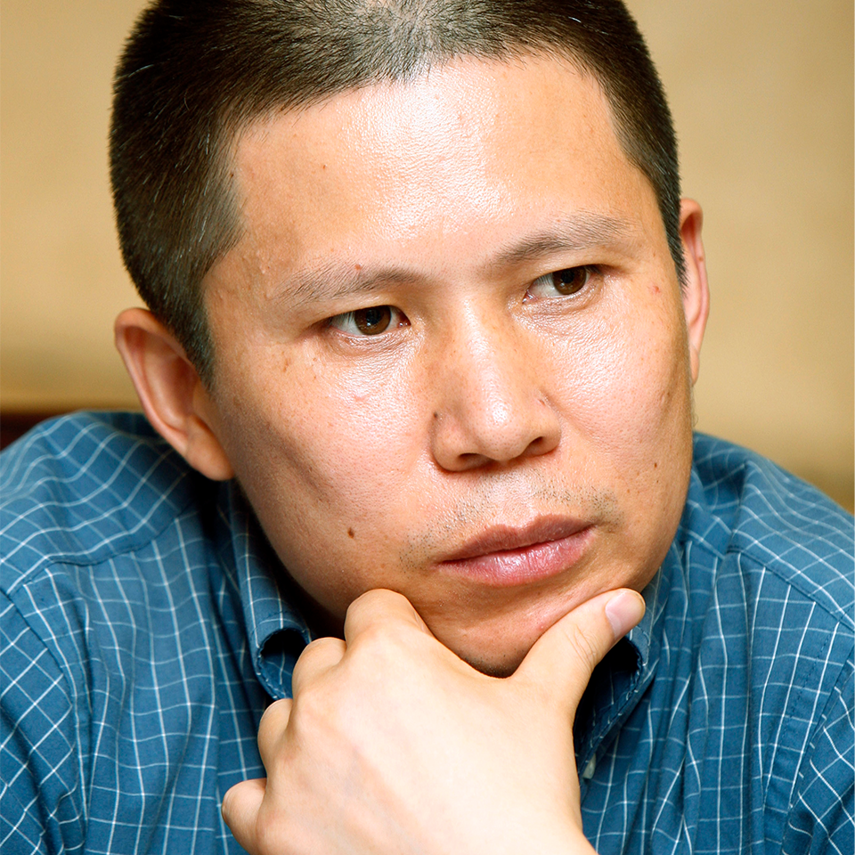 A man with short hair wearing a blue checkered shirt rests his chin on his hand, looking thoughtful and focused. The background is softly blurred.