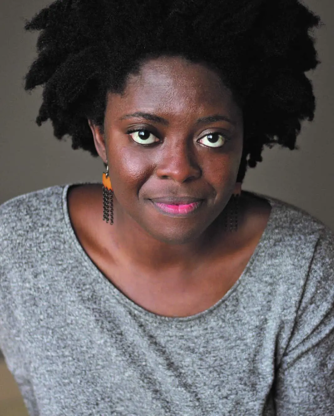 A woman with natural black hair and medium-dark skin wears a grey top and dangling earrings, looking slightly upward with a subtle smile against a neutral background.