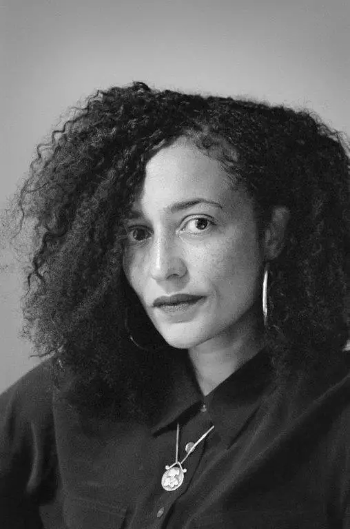 A woman with curly hair and hoop earrings gazes at the camera. She wears a dark button-up shirt and layered necklaces. The photo is black and white with a neutral background.