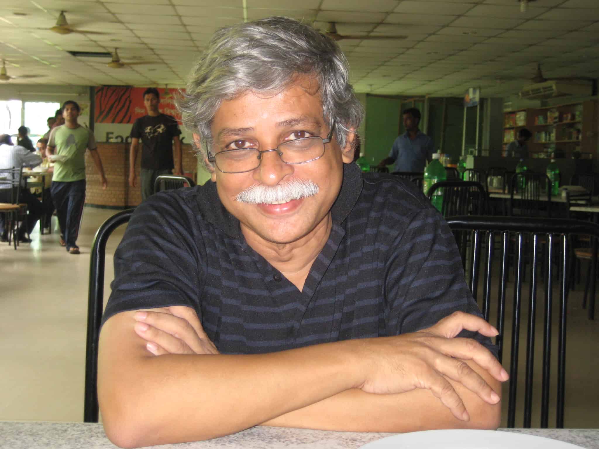 A smiling man with gray hair and glasses sits at a table in a cafeteria, arms crossed, wearing a black striped polo shirt. Other people and tables are visible in the background.
