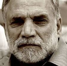 A close-up, black-and-white photo of an older man with a gray beard and mustache, showing deep facial lines, looking directly at the camera with a serious expression.