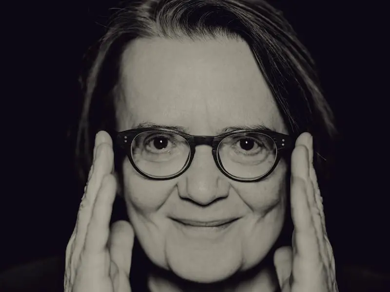 A person with glasses smiles softly, holding their hands beside their face. The image is in black and white, with a dark background, emphasizing their facial expression and features.