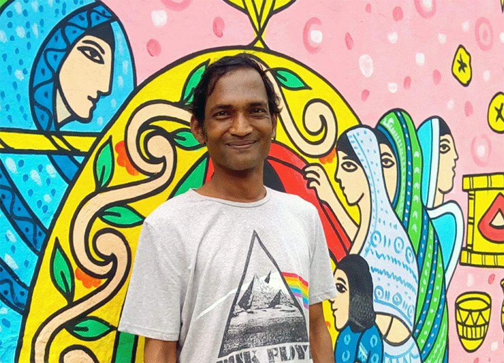 A man with dark hair smiles in front of a colorful mural featuring women in traditional clothing with intricate yellow, blue, and pink patterns. He wears a light gray T-shirt with a geometric design.