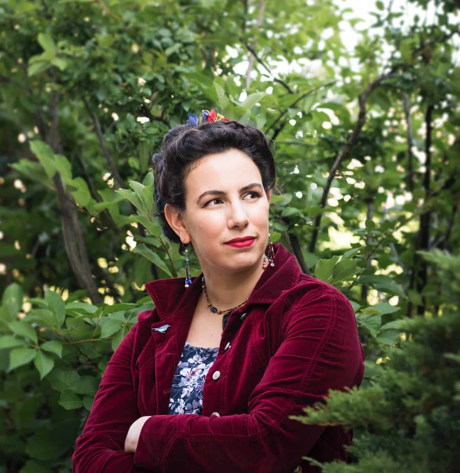 A woman with dark hair styled up, wearing a red velvet jacket and floral dress, stands with arms crossed in front of leafy green bushes, looking thoughtfully to the side, reminiscent of Azar Nafisi holding a pen.