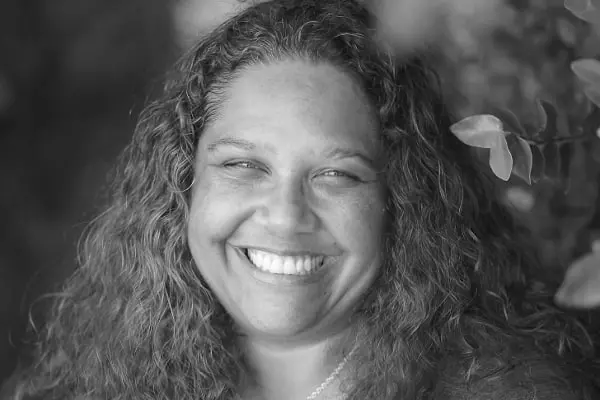 A woman with curly hair smiles broadly at the camera. The image is in black and white, and leafy branches are visible in the background.