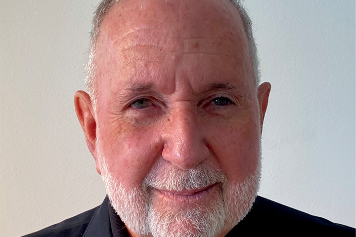 An older man with short gray hair and a neatly trimmed white beard smiles gently at the camera. He is wearing a black shirt and is in front of a plain, light-colored background.