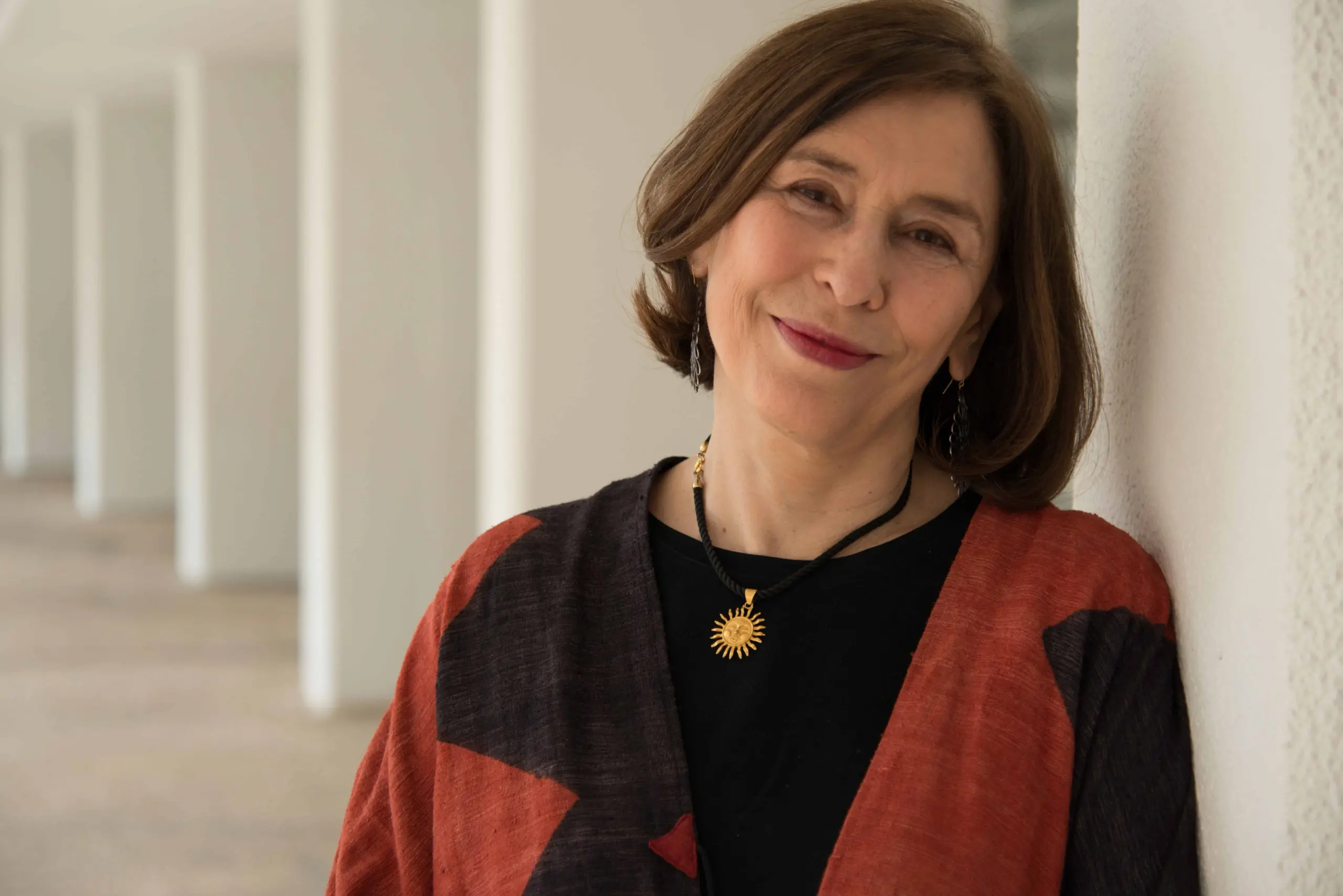 A woman with short brown hair, wearing a black top, a red and black patterned shawl, and a sun-shaped necklace—reminiscent of Azar Nafisi’s poise—smiles gently while leaning against a white wall in a bright, modern corridor.