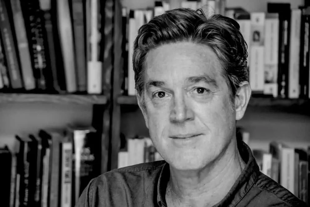 Black and white photo of a middle-aged man with short, wavy hair, wearing a collared shirt, sitting in front of bookshelves filled with books. He looks directly at the camera with a neutral expression.