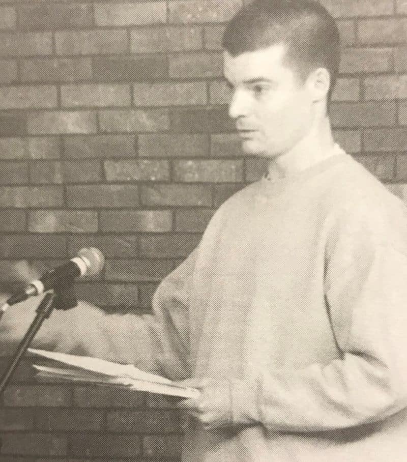 A young man in a sweatshirt stands in front of a brick wall, holding papers and speaking into a microphone on a stand—bringing to mind the candid style of B. Batchelor.