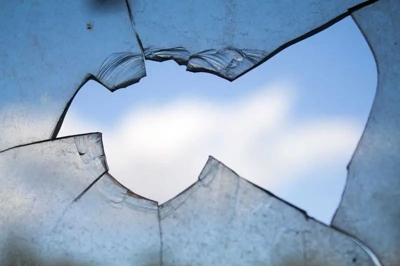 A close-up of a broken glass pane with a jagged hole in the center reveals a view of the blue sky and some white clouds outside. The edges of the glass are sharp and cracked.