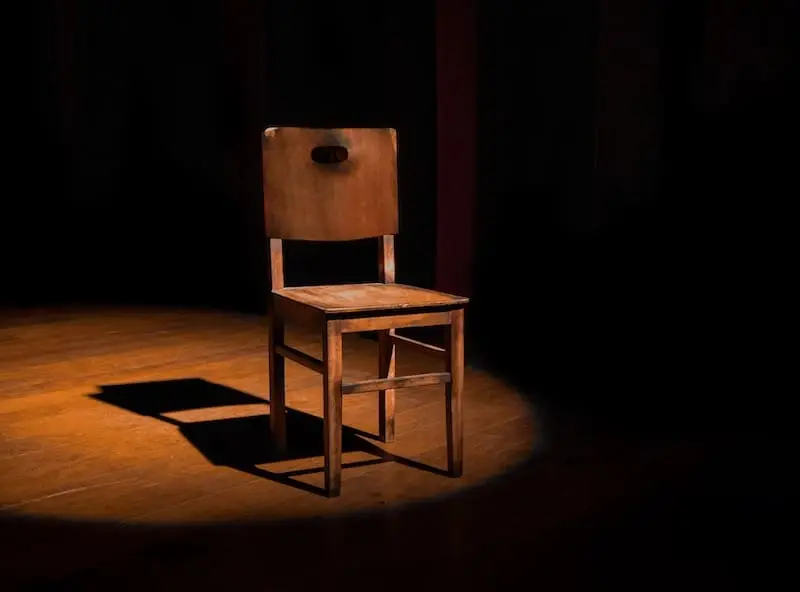 A single wooden chair is illuminated by a spotlight on a dark, empty stage, casting a shadow on the floor.