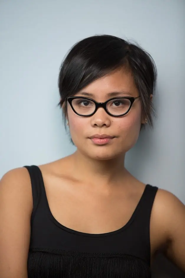 Cinelle Barnes, a woman with short dark hair and glasses wearing a sleeveless black top, stands against a plain light gray background, looking calmly at the camera.