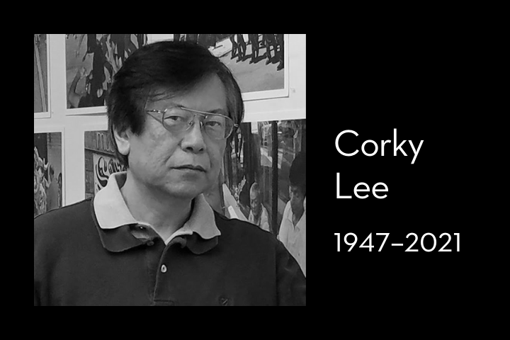 Black and white photo of a man with glasses standing in front of a wall with photographs, next to text reading “Corky Lee 1947–2021.”.