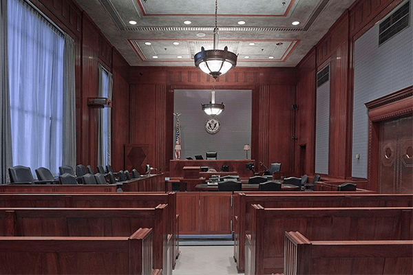 Empty courtroom with wooden benches, judge’s bench, chairs, and American flag. The room features large windows with blue curtains, chandeliers, and an official seal on the wall behind the judge’s seat.