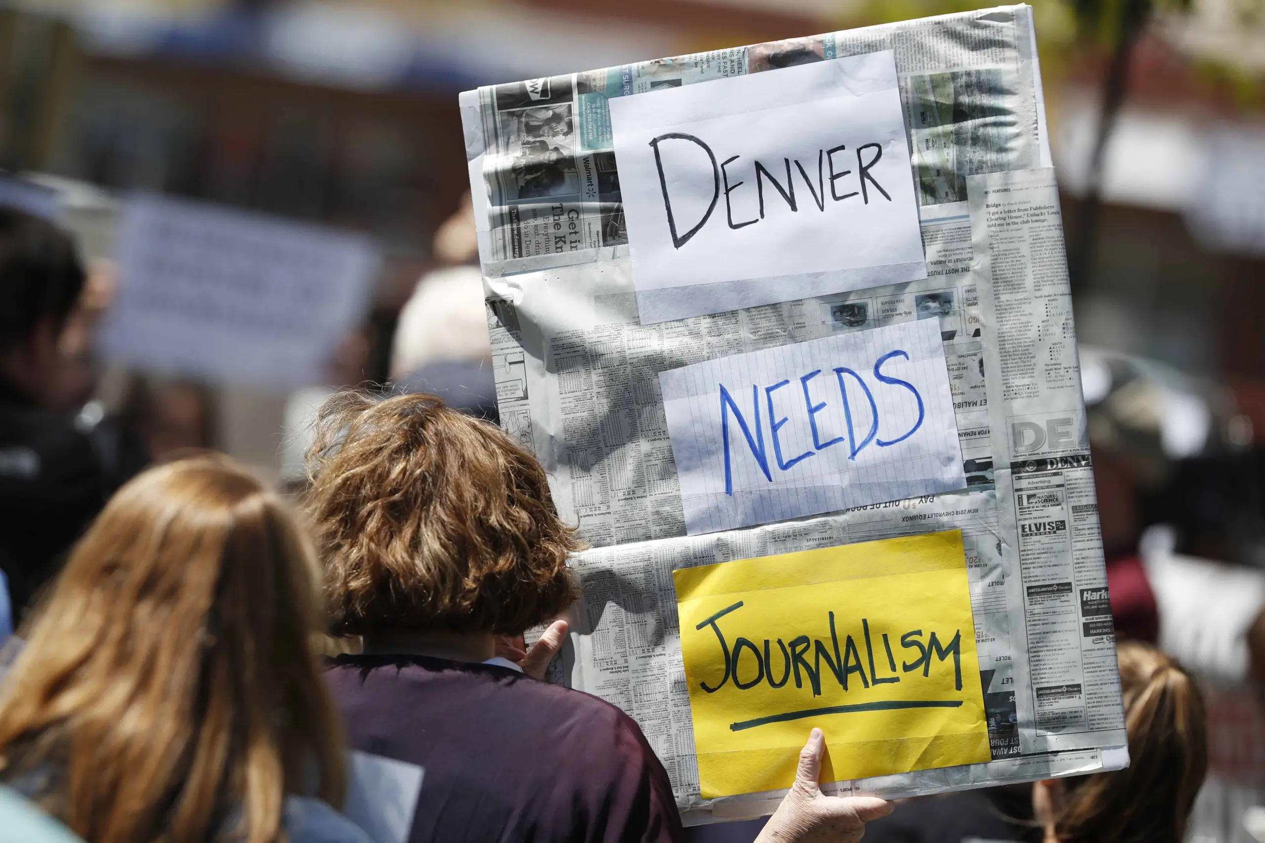 A person in a crowd holds a sign covered in newspaper pages with handwritten words: Denver Needs Journalism. Other people and blurred signs are visible in the background.