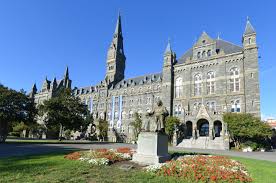 A large, historic stone building with a tall clock tower and pointed roofs stands behind a lawn with flower beds and statues under a clear blue sky.