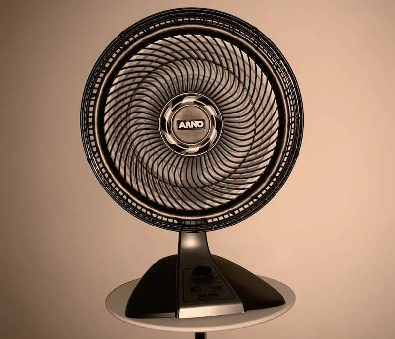A black and silver oscillating electric fan with a circular grille sits on a round white table against a beige background. The brand name ANNO is displayed in the center of the fan.